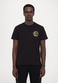 Tricou din bumbac negru cu guler rotund, mâneci scurte și un logo circular auriu pe pieptul stâng. Design simplu, de culoare solidă.