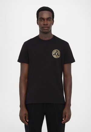 T-shirt en coton noir avec un col rond, des manches courtes et un logo circulaire doré sur la poitrine gauche. Design simple et de couleur unie.