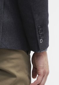 Zbliżenie ramienia osoby ubranego w ciemną, strukturalną marynarkę z czterema czarnymi guzikami na mankiecie oraz khaki spodniami na białym tle.