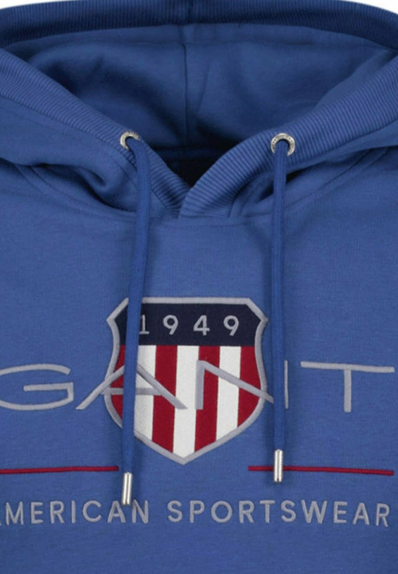 Sudadera azul con capucha y logo de escudo frontal que muestra "1949" y rayas rojas y blancas, texto "GANT" arriba y "American Sportswear" abajo.