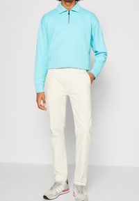Pullover azzurro chiaro con zip, pantaloni crema e sneakers grigie con accenti rossi. Tessuto liscio, vestibilità rilassata e design minimalista.
