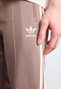 Bruna sportbyxor med strukturfinish, försedda med en vit Adidas-logotyp och krämfärgade sidosträck. Dragkedjeficka på vänster sida.