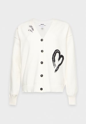 Un mannequin se tient devant un fond gris uni, portant un cardigan blanc à motif graphique, un jean bleu, une ceinture noire et des mocassins noirs.