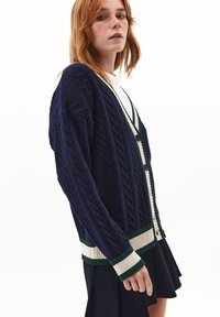 Cardigan de punto trenzado en navy con puños y dobladillo a rayas verdes y crema, con botones en el centro y un corte relajado.