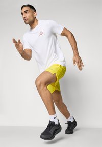 White athletic t-shirt with a small logo, paired with yellow shorts featuring side slits, completed with black sneakers and white socks.