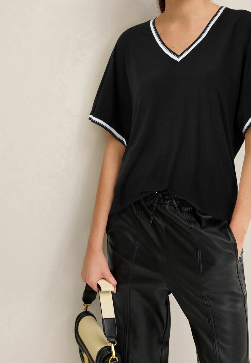 Femme portant un haut noir à col en V avec un bord blanc et un pantalon en cuir noir, tenant un sac à main beige et noir par la anse.