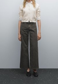 Cremfarbener Kabelstrick-Cardigan über einem weißen Hemd mit Kragen, kombiniert mit weit geschnittenen, dunkelblauen Jeans und schwarzen Slippers. Schlichtes und modernes Design.
