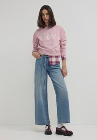 Felpa rosa con testo "Rosé Lovers", sovrapposta a una camicia a quadri. Jeans ampi blu e scarpe in pelle verniciata bordeaux.