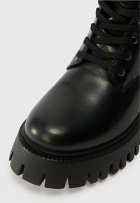 Μαύρο δερμάτινο μποτάκι μέχρι τον αστράγαλο με στρογγυλή μύτη, αντίθετη υφή στη σόλα και μαύρη κορδόνια. Αξιοσημείωτη παχιά, ανθεκτική σόλα με οδοντωτές άκρες.