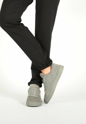 Persona vestida con pantalones negros y zapatillas de ante grises, de pie con un pie cruzado sobre el otro, sobre un fondo blanco.