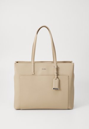 Sac fourre-tout en cuir beige avec deux bretelles, une poche avant et une petite étiquette attachée avec des détails métalliques.