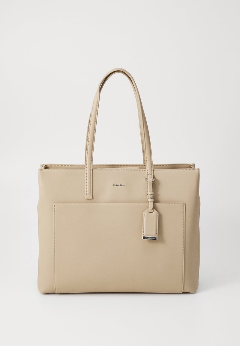 Sac fourre-tout en cuir beige avec deux bretelles, une poche avant et une petite étiquette attachée avec des détails métalliques.