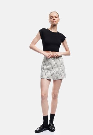 Vrouw staand, draagt een zwart crop top, een rok met hoge taille en patroon, zwarte sokken en zwarte loafers op een witte achtergrond.