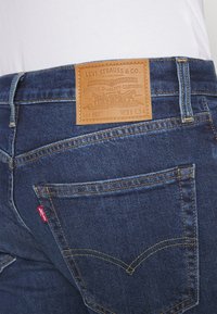 Tmavě modré džíny z denimu s koženou nášivkou se nápisem "Levi Strauss & Co." prošitou na zadní straně. Kapsy zdobí žluté švy.