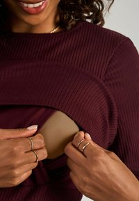 Bordeaux ribbed top dengan bagian yang dipotong yang sedang ditarik. Tangan menunjukkan cincin emas, menyoroti tekstur halus dan desain yang pas.