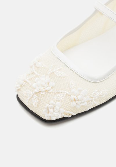 Chaussures Mary Jane en maille blanche avec des embellissements floraux, une surface texturée et une semelle noire plate ; comprennent une sangle blanche sur le coup de pied.