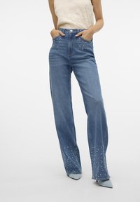 Jeans in denim blu chiaro con un design a gamba larga, impreziositi da borchie metalliche sulle tasche frontali e un dettaglio di orlo con borchie.