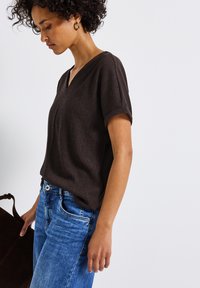 Brun, strukturerad kortärmad topp med V-ringning, tillsammans med blå denimjeans. Modellen håller en mörkbrun väska.