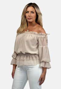 Bluzka w kolorze jasnobeżowym, bez ramiączek, z falbaniastymi rękawami i marszczonym w pasie, wykonana z fakturowanej, zwiewnej tkaniny. Noszona z białymi spodniami.