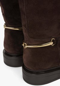 Donkerbruine suede laarzen met een gladde textuur, hoogte tot de enkel, en een goudkleurig hardwareaccent aan de achterkant. Dikke zwarte plateauzool.