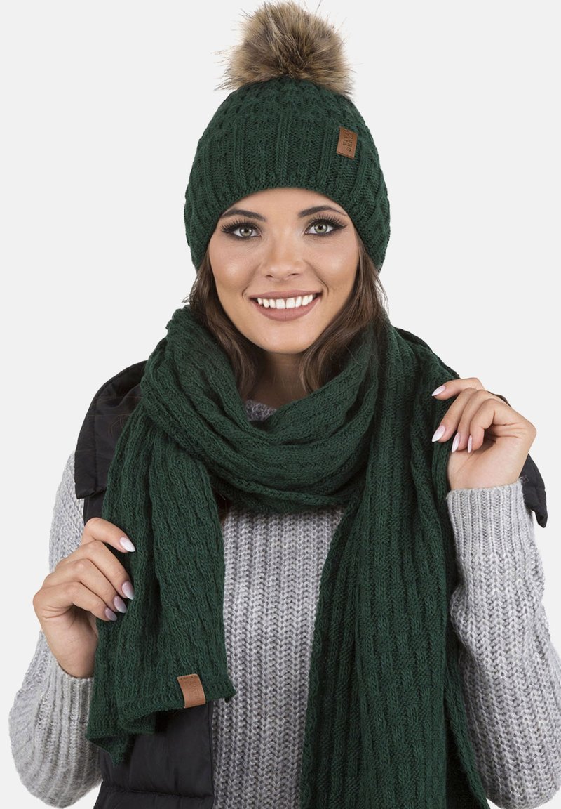 Zöld kötött sapka műszőrme pom-pommal, egy matching textúrájú sállal párosítva. A sapkán egy kis bőrcímke látható.