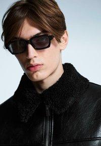 Crna kožna jakna s teksturiranim crnim krznom ovratnikom, uparen s odvažnim crnim sunčanim naočalama s geometrijskim dizajnom.