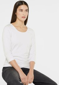 Weißes Langarmhemd mit Rundhalsausschnitt. Weicher Baumwollstoff. Kombiniert mit dunkelgrauen Jeans, Modell sitzt mit Händen auf den Oberschenkeln.