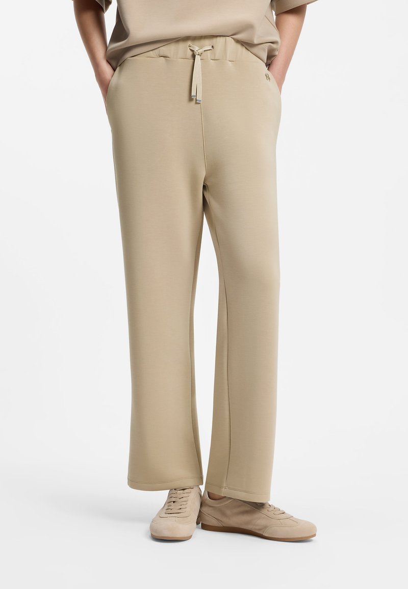 Personne portant un pantalon beige à jambes larges avec une taille à cordon et des baskets beige assorties, mains dans les poches, sur un fond blanc uni.