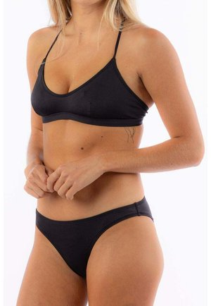 Frau in einem schwarzen Bikini mit dünnen Trägern, die vor einem weißen Hintergrund steht, die Hände nahe ihrer Taille gefaltet.