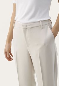 Pantaloni beige chiaro realizzati in tessuto testurizzato, con vestibilità dritta, davanti piatto e tasche laterali. Indossati con un top bianco con righe.