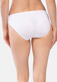 Donna che indossa una semplice mutandina bianca di cotone, vista da dietro, su uno sfondo chiaro e neutro.