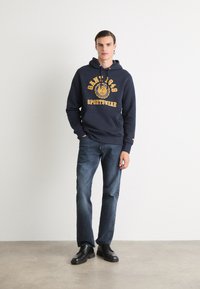 Navy hooded sweatshirt with yellow text and logo, paired with dark blue jeans and black shoes. Smooth fabric, relaxed fit, front pocket.