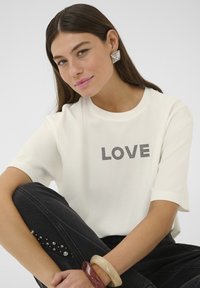 Bela kratka majica s kratkimi rokavi, na kateri je natisnjen napis "LOVE" v črni barvi. Model nosi črne kavbojke z okraski in velikim zapestnim obročem.