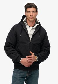 Zwarte capuchonjack met een voorzichtige rits, geribbelde manchetten en zakken; gedragen over een witte overhemd met kraag, gecombineerd met blauwe spijkerbroeken.