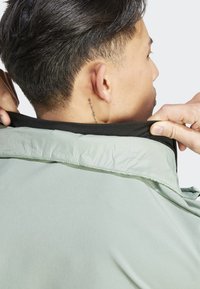Chaqueta verde claro con una textura suave y semi-brillante, que presenta un collar negro y costuras detalladas. Tatuaje visible en la parte posterior del cuello.
