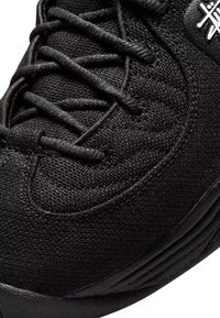 Baskets noires avec texture en tissu tissé, lacets noirs et un petit logo blanc près de la cheville, montrant un gros plan de la partie supérieure avant et du bord de la semelle.