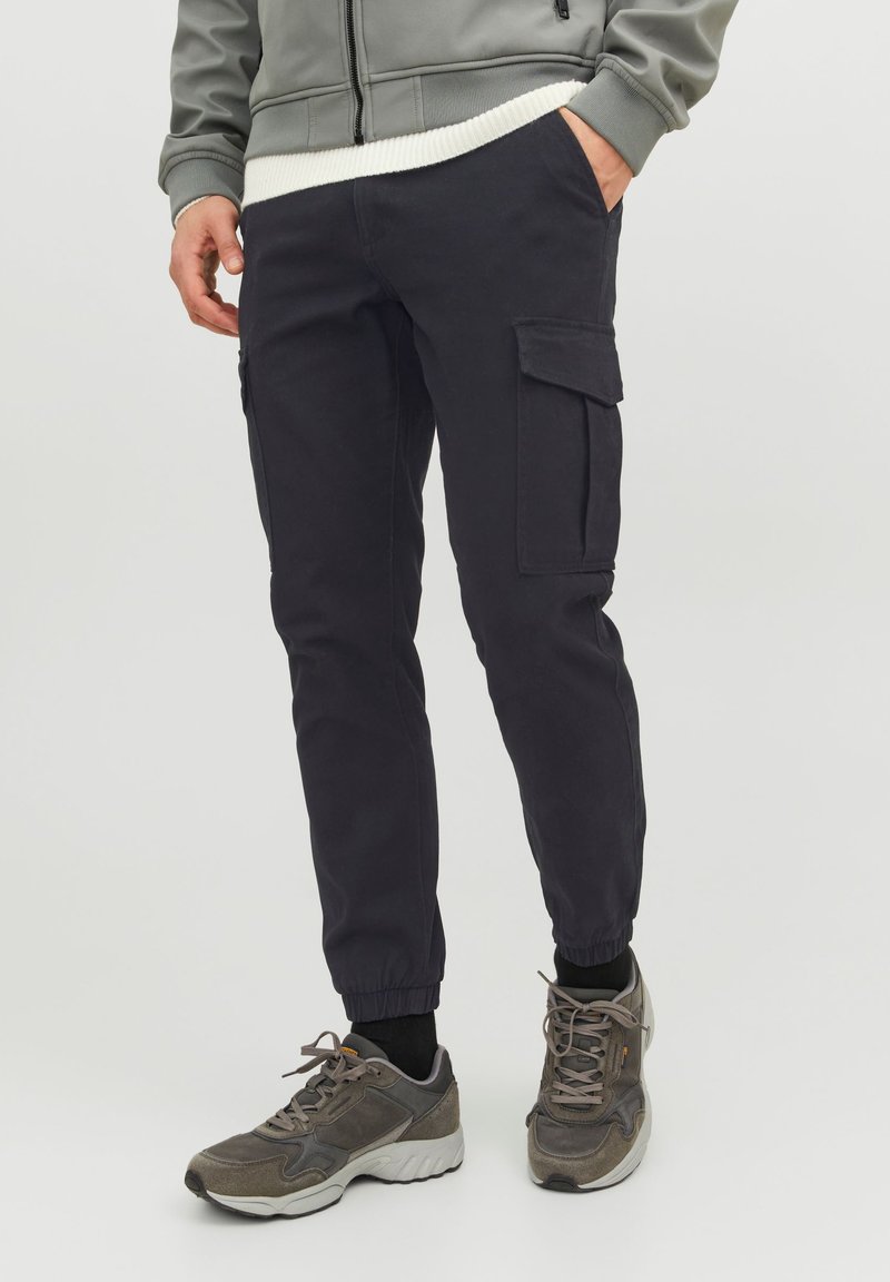 Homme portant un pantalon cargo jogger noir avec des poignets élastiques et des baskets grises, debout avec une main dans la poche.