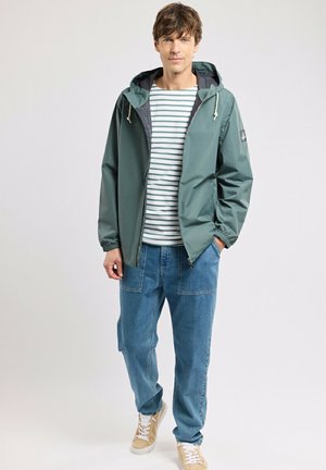 Homme portant une veste à capuche verte par-dessus une chemise blanche et verte à rayures, un jean bleu avec de grandes poches et des baskets beige sur fond clair.