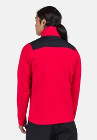 Červená dlouhá bunda s dlouhým rukávem a černým ramenním panelem, s vysokým límcem a hladkým povrchem. Design je přiléhavý a minimalistický.