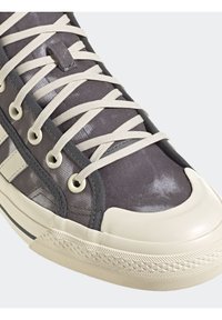 Teniska se šedým textilním svrškem, krémovou gumovou špičkou a krémovými tkaničkami. Vyznačuje se texturovanými detaily a prošitými akcenty podél stran.