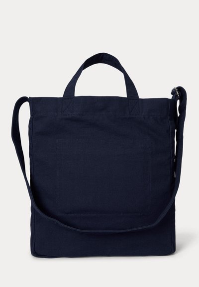 Τσάντα ώμου από καμβά σε ναυτικό μπλε χρώμα με κοντά χερούλια, μπροστινή τσέπη και ρυθμιζόμενο λουρί ώμου, σε απλό φόντο.