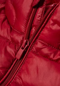 Materiál červené péřové bundy s vystlanou texturou, s červeným zipem a odpovídajícím nylonovým jezdcem podél přední části.