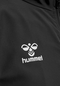 Sort jakke med en glat tekstur. Viser et hvidt logo med et bi-design og ordet "hummel" nederst.