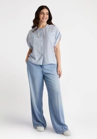 Maglietta azzurro chiaro a maniche corte con bottone e tasche, abbinata a pantaloni in denim chiaro a vita alta e morbidi. Texture liscia, vestibilità comoda e dettaglio con cordino.