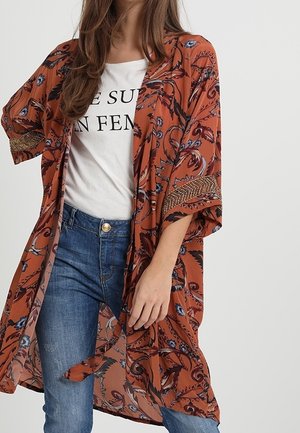 Kobieta ubrana w rdzawy, kwiatowy kimono na białym koszulce z nadrukiem i niebieskich jeansach, stojąca na jednolitym jasnym tle.
