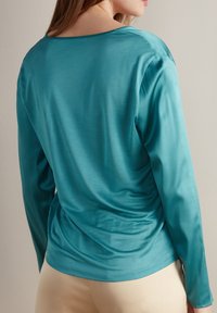 Kvinna i en teal långärmad silktopp och beige byxor, sedd bakifrån mot en enfärgad ljus vägg.