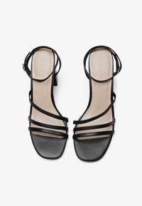 Black heeled sandals with a smooth leather upper, featuring multiple thin straps and a square toe. Light-colored insole, adjustable ankle strap.