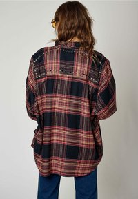 Camicia a scacchi nera e borgogna oversize con maniche corte, caratterizzata da dettagli borchiati lungo la cucitura della spalla e il giogo posteriore.