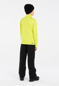 Maglietta a maniche lunghe e colletto alto di un vivace colore giallo, abbinata a pantaloni neri. L'outfit presenta una texture liscia e un branding minimale sulla maglietta.