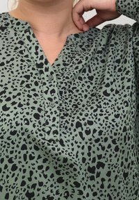 Top verde con un patrón de leopardo negro, que presenta un escote en V y una tapeta de botones. El material parece ligero y texturizado.